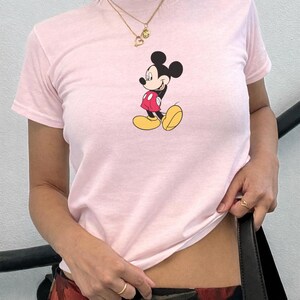 May include: A pale pink t-shirt with a Mickey Mouse graphic. The cartoon character is shown in his iconic outfit: black ears, red jacket, yellow shoes, and white gloves. The top is short-sleeved.