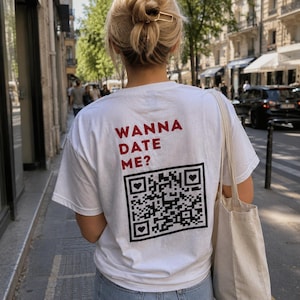 Può includere: T-shirt bianco con la scritta "WANNA DATE ME?" in lettere rosse. Sotto il testo c'è un codice QR in bianco e nero con simboli a forma di cuore. La persona indossa jeans blu e una borsa tote beige.