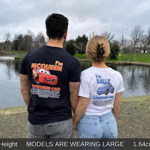 Può includere: Due persone indossano magliette abbinate con grafiche del film Cars. Una maglietta è nera con "I'm McQueen" e un'immagine di Saetta McQueen rossa. L'altra è bianca con "I'm Sally" e un'immagine di Sally Carrera blu. Sono in piedi vicino a uno specchio d'acqua.