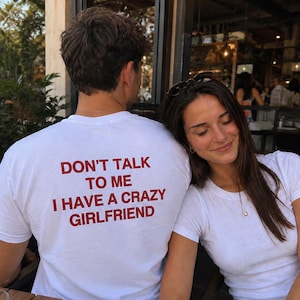 Può includere: Una t-shirt bianca a maniche corte con testo rosso sul retro che recita: DON'T TALK TO ME I HAVE A CRAZY GIRLFRIEND. La maglia è realizzata in tessuto bianco semplice con scollo rotondo e una vestibilità standard, mostrando il messaggio.