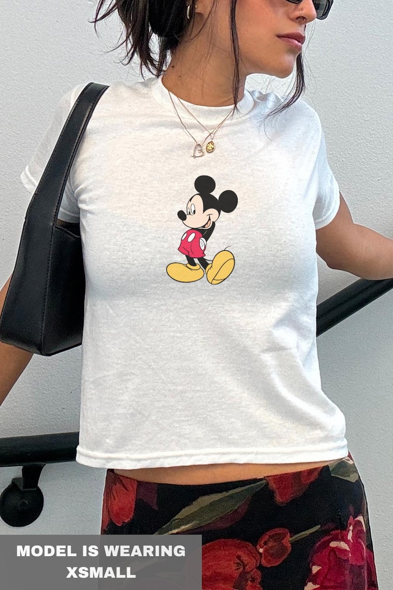 May include: White t-shirt with a Mickey Mouse graphic in a red jacket, yellow shoes, and black shorts. The outfit includes a black bag and a floral skirt. The text "MODEL IS WEARING XSMALL" is visible.