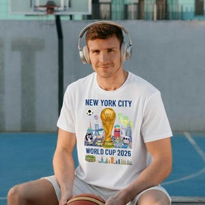 May include: White t-shirt with a colorful graphic featuring the text "NEW YORK CITY WORLD CUP 2026" and a gold trophy. The design includes illustrations of New York City landmarks. A person is wearing the shirt and holding a basketball.