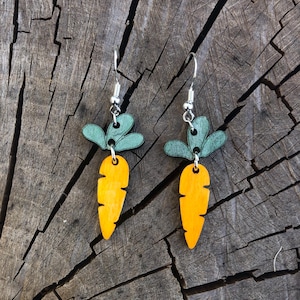 Può includere: Un paio di orecchini in legno a forma di carota, con ganci color argento. Gli orecchini presentano carote arancioni con cime verdi. Sono esposti su una superficie di legno rustica e invecchiata.