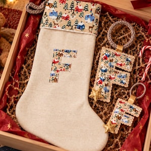 May include: A wooden Christmas Eve Box containing a beige stocking with a winter village print cuff and a letter F applique, two matching letter F ornaments with gold stars, and a candy cane, all nestled on red tissue paper and brown filler.
