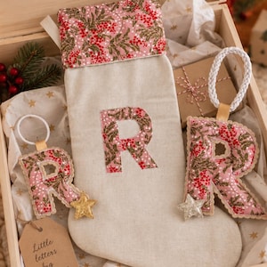 May include: A wooden Christmas Eve box containing a beige stocking with a pink floral cuff and a large letter R, two matching letter R ornaments with white braided loops and gold star accents, and a brown tag labelled Little letters big memories.