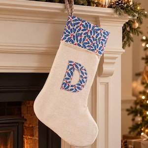 May include: A cream-coloured fabric Christmas stocking featuring a cuff and a large letter D applique, both decorated with a blue holly leaf and red berry pattern. The stocking includes a braided loop at the top for hanging purposes.