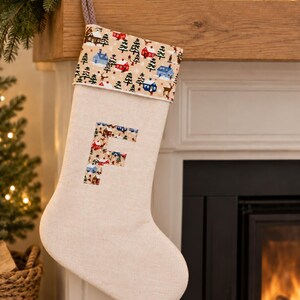 May include: A beige fabric Christmas stocking featuring a cuff with a winter village pattern of houses, trees, and deer. The letter F is appliqued on the centre of the stocking in the same patterned fabric. A braided loop is attached to the top corner.