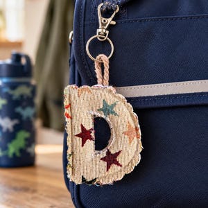 May include: A fabric letter D keyring featuring a beige textured background with a pattern of small red, green, and orange stars. The letter has frayed edges and is attached to a silver metal lobster clasp by a short, thick, tan-coloured rope loop.