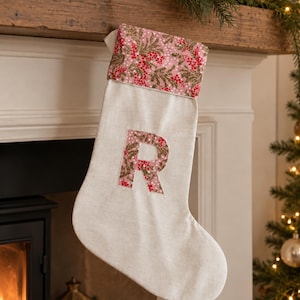 May include: A cream-coloured fabric Christmas stocking featuring a cuff and a large letter R applique. Both the cuff and the letter are decorated with a repeating pattern of pink flowers, red berries, and green foliage on a light pink background.