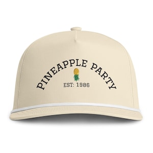 May include: A cream-colored baseball cap featuring a white cord trim along the base of the crown. The front displays the black text PINEAPPLE PARTY in an arched layout, with a small yellow and green pineapple graphic and the text EST: 1986.