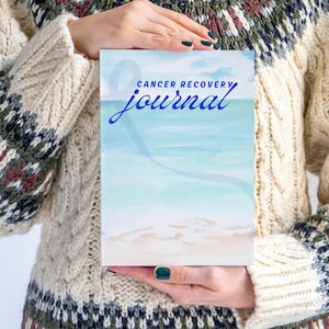May include: A journal with the words "Cancer Recovery Journal" in blue script. The cover features a watercolor beach scene with a blue sky and white sand. The journal is held by a person wearing a cream-colored sweater with patterned sleeves.