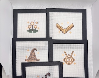 May include: Five square needlepoint canvases with black borders, each featuring a small pixel-art style design: a glowing plant, golden wings, a brown pointed hat, a golden hourglass pendant, and a pink umbrella, all accented with small yellow stars.