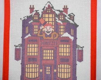 May include: A cross-stitch embroidery on white fabric with a bright orange border, depicting a multi-story purple and red building with yellow windows, snow on the roof, and a figure waving from an upper window, with the text Wheezes visible.
