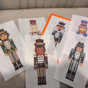 May include: Six rectangular needlepoint canvases featuring colorful nutcracker designs. The figures include a bearded man in green, a horse, a tiger, a bird, a brown animal with tusks, and a dark-haired figure in a black and green uniform.