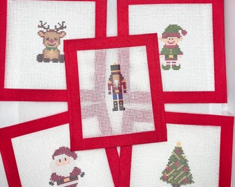 May include: A set of five square needlepoint canvases with red fabric borders. Each white canvas features a different cross-stitched holiday design: a reindeer, an elf, a nutcracker, Santa Claus, and a decorated Christmas tree.