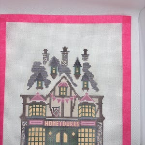 May include: A needlepoint canvas featuring a cross-stitch design of a building with the text HONEYDUKES. The design includes a dark green storefront, yellow windows, and a roof with chimneys, all framed by a bright pink border on white mesh fabric.
