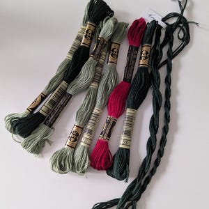 May include: A selection of embroidery threads in shades of green and red. The threads are bundled and labelled with numbers. Some threads are loose and twisted. The image is taken on a white surface.