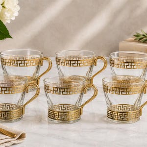 May include: Six clear glass teacups with gold-colored handles and decorative gold Greek key patterns. The teacups are arranged on a white surface, with a gold spoon and a vase of white flowers in the background.