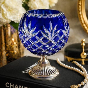 May include: A cobalt blue crystal glass bowl with a silver-colored pedestal base. The bowl features a diamond and leaf-shaped cut pattern. The glass bowl is sitting on a black surface.