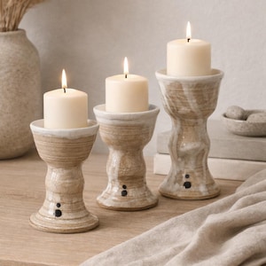 May include: Three ceramic candle holders with lit, ivory-colored candles. The holders are beige with a white rim and black accents. The candles are cylindrical and have a soft glow. The holders are on a wooden surface.