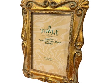 May include: A gold-toned picture frame with ornate, swirling scrollwork edges and a beaded inner border. The insert displays the text Towle Silversmiths Since 1690, Signature Frame and Frame Album Collection, and Genuine Ceramic Four By Six.