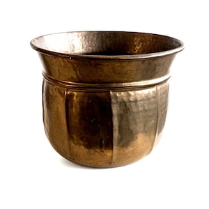 May include: A round, metallic planter with a warm, bronze-toned finish. The exterior surface features a distinct hammered texture and vertical indentations that create a paneled appearance. The rim is wide and slightly flared, with a smooth, rounded edge.