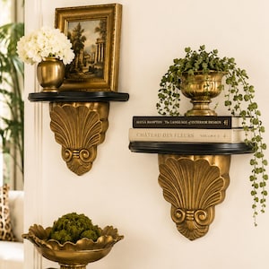 May include: Two wall-mounted shelves with black surfaces and gold-colored, shell-shaped brackets. The left shelf holds a gold vase with white flowers and a framed landscape painting. The right shelf holds a gold planter with trailing greenery and two books.