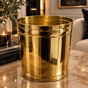 May include: A cylindrical wastebasket with a polished, reflective gold-tone finish. The container features a smooth, metallic surface with subtle horizontal ridges near the top and bottom rims, creating a clean, uniform, and highly reflective appearance.