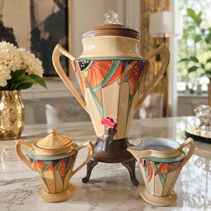 May include: A ceramic beverage service set featuring a large urn with a spigot, a lidded sugar bowl, and a creamer. Each piece has a cream-colored base with geometric patterns in orange, teal, and black, and includes two handles and a pedestal base.