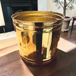May include: A large, cylindrical, polished brass wastebasket featuring a reflective, metallic gold finish and vertical ribbed detailing along the exterior body. The rim is smooth and circular, with a small red circular sticker visible inside.