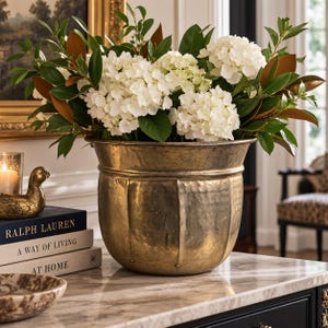 May include: A large, hammered brass planter with a rounded, fluted body and a wide rim, filled with a lush arrangement of white hydrangea blossoms and deep green magnolia leaves, creating a substantial decorative centerpiece for a home interior.