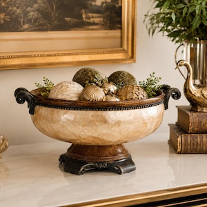 May include: A decorative bowl with a cream-colored, textured shell-like finish, featuring dark brown scrolled handles and a matching pedestal base. The bowl is filled with assorted spherical ornaments in moss green, gold, and cream tones.