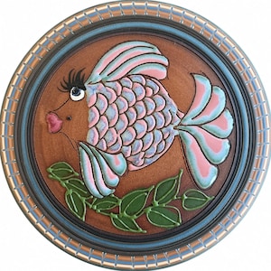May include: A round ceramic plate with a brown background featuring a stylized fish with pink and light blue scales, a pink mouth, and long eyelashes. Below the fish is a green leafy vine. The plate has a decorative blue and brown rim.