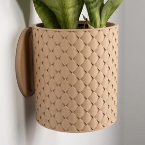 May include: A beige wall-mounted plant pot with a woven texture, containing a green plant. The pot has a curved handle for hanging. The plant pot is suitable for indoor or outdoor use.