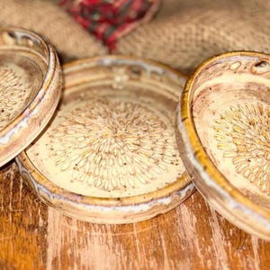 May include: Three round ceramic dishes with raised edges, featuring a cream-colored glaze and a central stamped floral pattern in brown. The exterior rims show a blend of tan, brown, and blue tones, with a textured finish on the interior base.