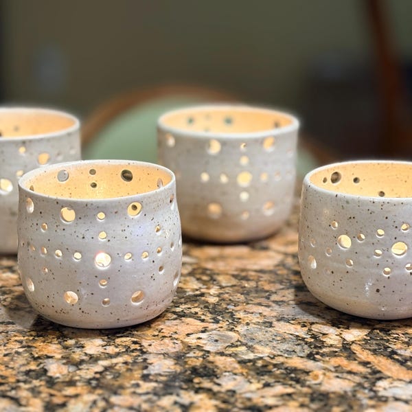 May include: A set of four off-white ceramic candle holders with a speckled finish and various circular cutouts throughout the body. Each cylindrical vessel features a smooth rim and a hollow interior that allows light to pass through the openings.