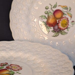 May include: White ceramic plates with a scalloped edge and a raised dot pattern. Each plate features a colorful fruit and floral design in the center, including pears, peaches, and flowers. The plates are stacked, with the top plate partially obscuring the one below.