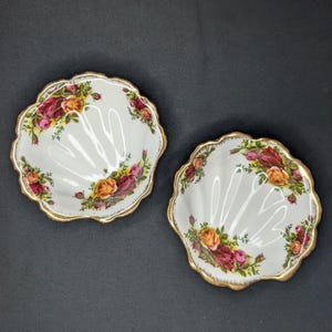 Puede incluir: Dos platos blancos en forma de concha con ribete dorado y estampado floral de rosas. Las rosas son en tonos rojos, rosas y amarillos. Los platos están sobre una superficie gris oscuro.