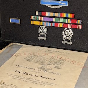 May include: A display of military medals and ribbons, including a blue and silver badge with a rifle, and a certificate from The Wall of Liberty Foundation. The certificate is for PFC Warren L. Anderson.