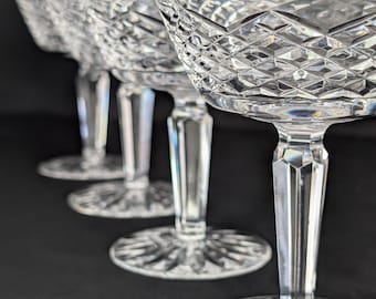 May include: Four clear crystal champagne coupe glasses. The glasses have a diamond-cut pattern on the bowl and a faceted stem. The base of each glass is round with a starburst design. The glasses are arranged in a row against a dark background.
