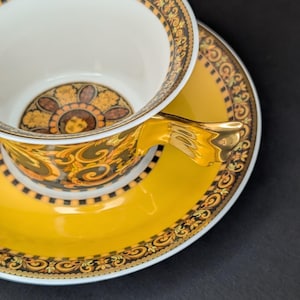 Könnte beinhalten: Ein gelbes Tee-Set mit aufwendigen Gold- und Schwarzdetails. Die Tasse und Untertasse haben einen dekorativen Rand und ein zentrales Design. Der Griff der Tasse ist goldfarben. Das Set ist wahrscheinlich aus Porzellan.