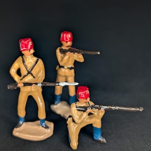 Puede incluir: Tres soldados de juguete en uniformes beige, sombreros rojos y botas azules, sosteniendo rifles. Las figuras están pintadas con detalles negros y se alzan sobre bases beige sobre un fondo oscuro. Los soldados están en varias poses.