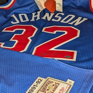 Puede incluir: Una camiseta de baloncesto azul con el nombre "JOHNSON" y el número "32" en rojo. La camiseta tiene ribetes blancos y un parche "Hardwood Classics". Hay etiquetas adjuntas. La camiseta es de la temporada 1991-92.