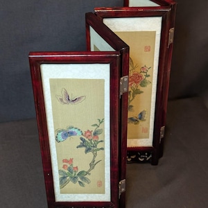 May include: A four-panel folding screen with a dark red wooden frame and delicate floral and butterfly paintings on each panel. The screen is decorated with intricate carvings on the bottom.