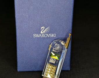 Może przedstawiać: Przezroczysta kryształowa figurka w kształcie telefonu komórkowego z klapką w stylu vintage ze złotymi akcentami, zielonym prostokątnym kryształowym ekranem i złotą klawiaturą. Figurka znajduje się przed ciemnoniebieskim prostokątnym pudełkiem z logo Swarovski.