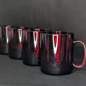 Op de afbeelding: Vier donkerpaarse glazen mokken met afgeronde handvatten zijn in een rij gerangschikt. De mokken hebben een cilindrische vorm en een glanzende afwerking. De achtergrond is donkergrijs.