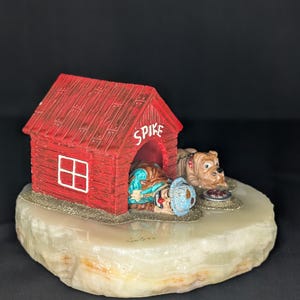 May include: A decorative figurine featuring a red doghouse with the name SPIKE written above the entrance. A small figure wearing a blue hat rests inside the house, while a brown bulldog lies next to a small food bowl on a textured stone base.