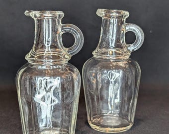 Peut inclure: Deux bouteilles en verre transparent avec une surface texturée et une poignée. Les bouteilles ont un corps arrondi et un col étroit.