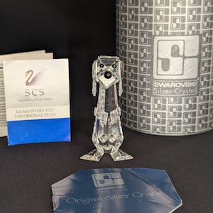 Puede incluir: Una figura de perro de cristal transparente con un cuerpo largo y orejas grandes. La figura está sobre una superficie negra, con una caja y papeles en el fondo. La caja dice "SWAROVSKI Silver Crystal".