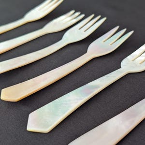 May include: Six mother-of-pearl forks with three tines and angled handles. The forks have iridescent colours of white, cream, and pale green. They are arranged diagonally against a dark grey background, showcasing their elegant design.